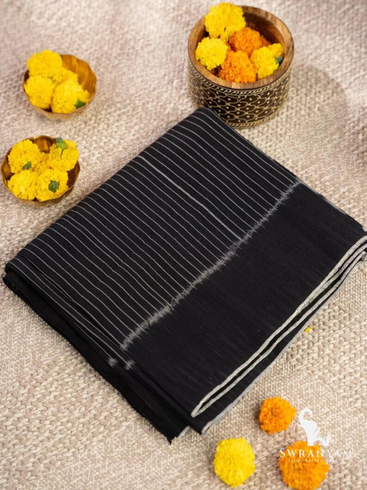 Folded black Pochampally ikat cotton saree with fine white stripes and plain black pallu beside marigold flowers