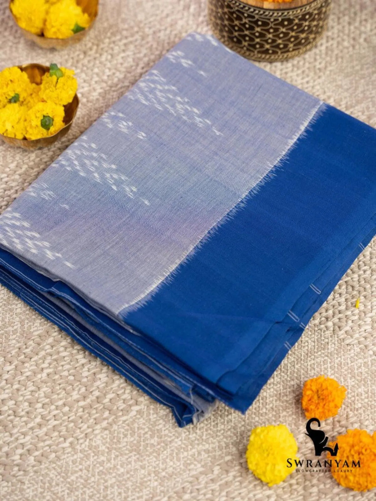 Folded sky blue Pochampally ikat cotton saree with royal blue border and white ikat motifs beside marigold flowers