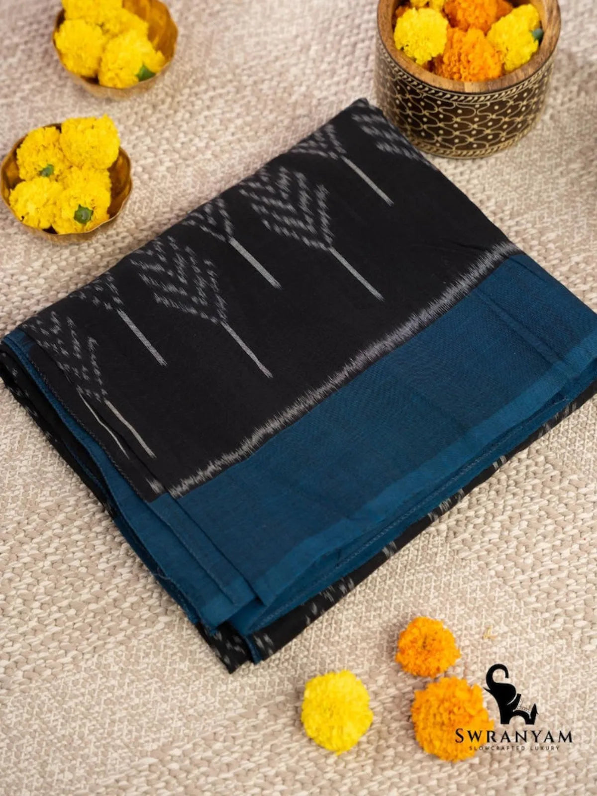 Folded black Pochampally ikat cotton saree with teal blue border and white leaf ikat motifs beside marigold flowers