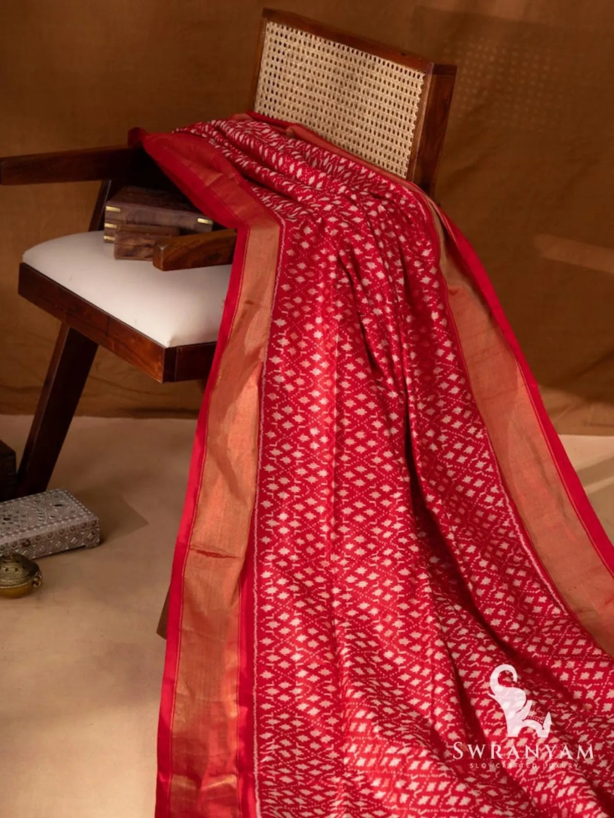 Red silk saree with all-over white single ikat geometric motifs and gold zari border draped on a wooden chair