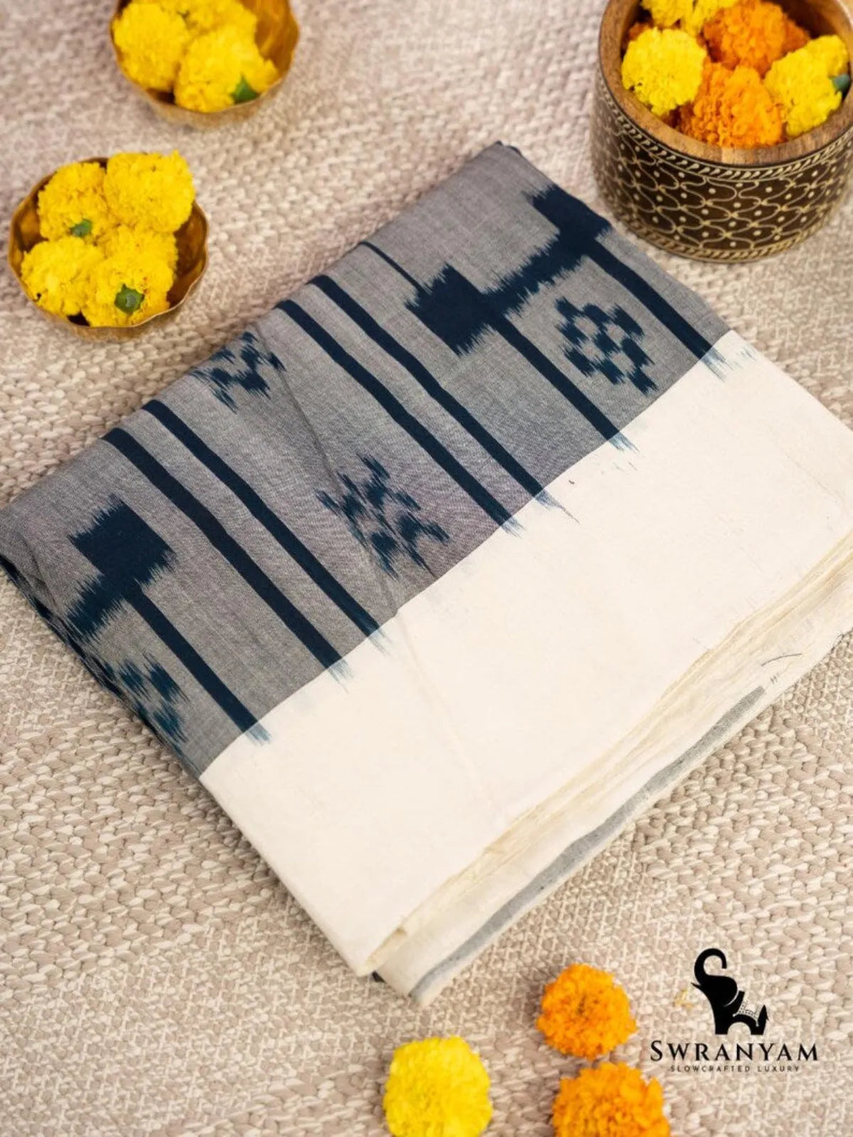 Folded grey Pochampally ikat cotton saree with indigo blue motifs and ivory border beside marigold flowers