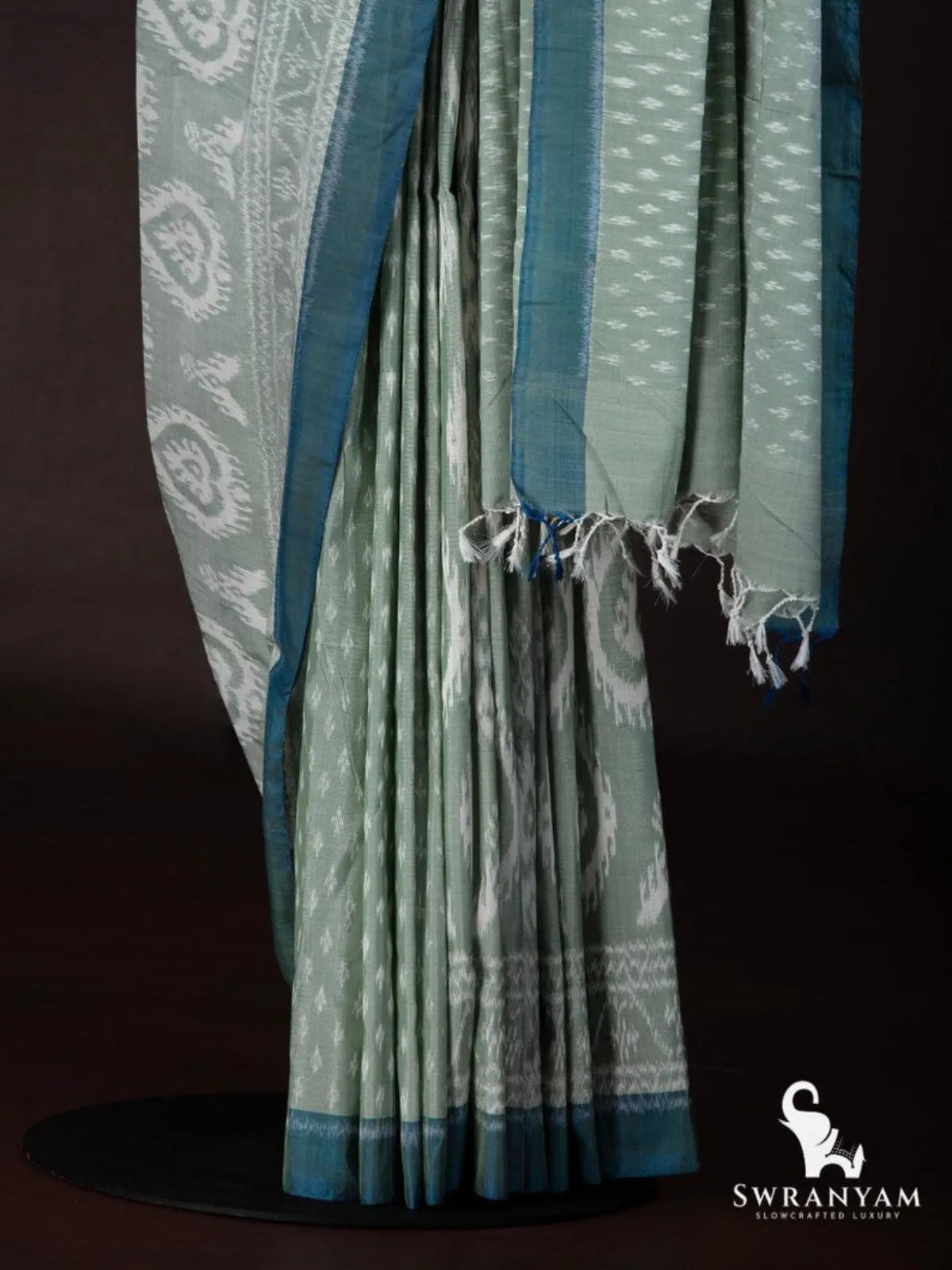 Pleats and tasselled pallu of sage green ikat silk cotton saree with small white butti motifs and teal border