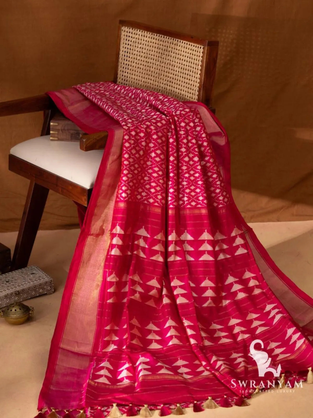 Pink silk saree with all-over white geometric ikat motifs and wide gold zari borders draped on a wooden chair