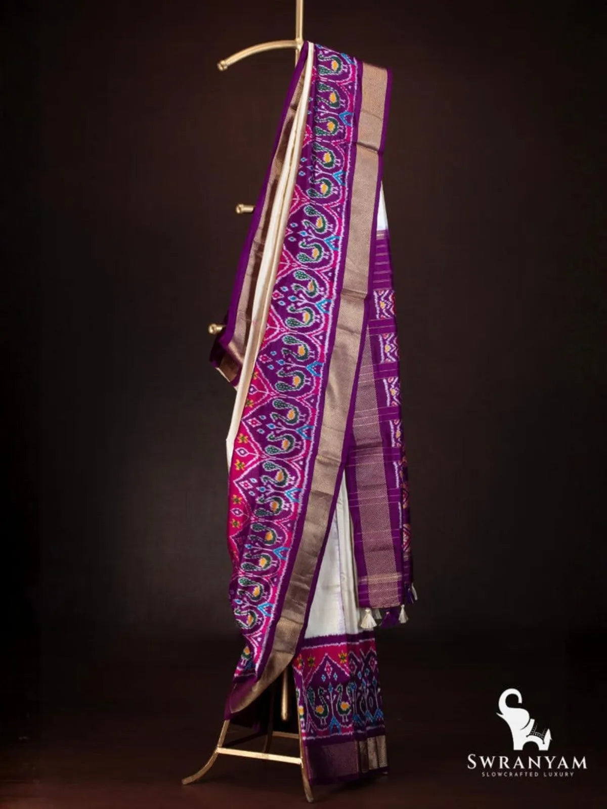 Side view of ivory ikat silk saree with royal purple and pink peacock ikat border and zari pallu on a brass stand