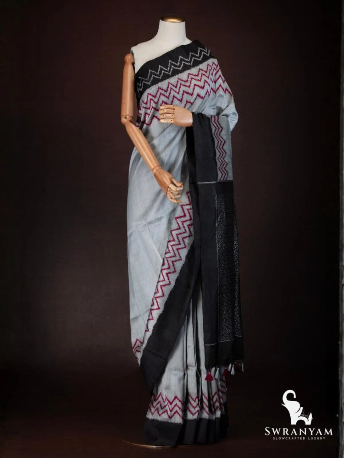Side view of charcoal grey tussar silk saree with red zigzag ikat border, black pallu and tassels on mannequin